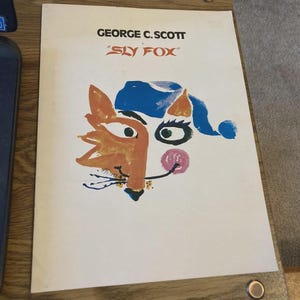 Puede incluir: Un póster vintage con el texto "GEORGE C. SCOTT" y "SLY FOX". El póster presenta una ilustración de un zorro en naranja, azul y rosa. El póster está sobre una superficie de madera.