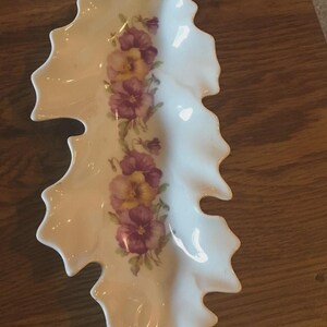 May include: White ceramic leaf-shaped dish with a gold-colored tip. The dish is decorated with a floral pattern of purple and yellow pansies. The dish is likely used for serving or display.
