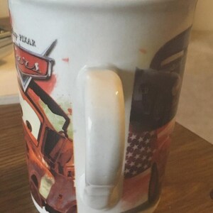 May include: White ceramic mug with a design from the Disney Pixar Cars film. The mug has a large handle and features the Cars logo and character images. The mug is cylindrical in shape and suitable for hot beverages.