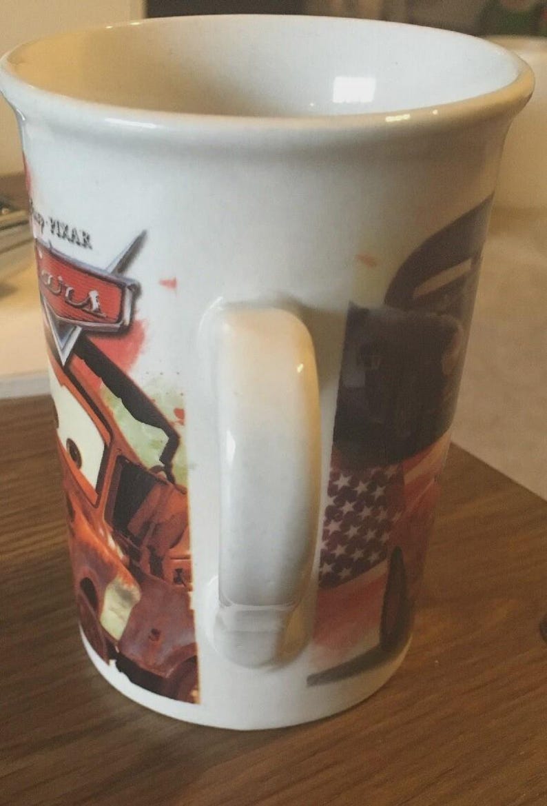 May include: White ceramic mug with a design from the Disney Pixar Cars film. The mug has a large handle and features the Cars logo and character images. The mug is cylindrical in shape and suitable for hot beverages.