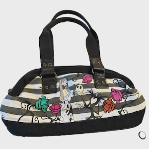 Puede incluir: Bolso de mano a rayas blancas y negras con personajes de The Nightmare Before Christmas. Tiene asas y ribetes negros, con cierre de cremallera. El dise&ntilde;o incluye a Sally, Jack Skellington y rosas de colores.