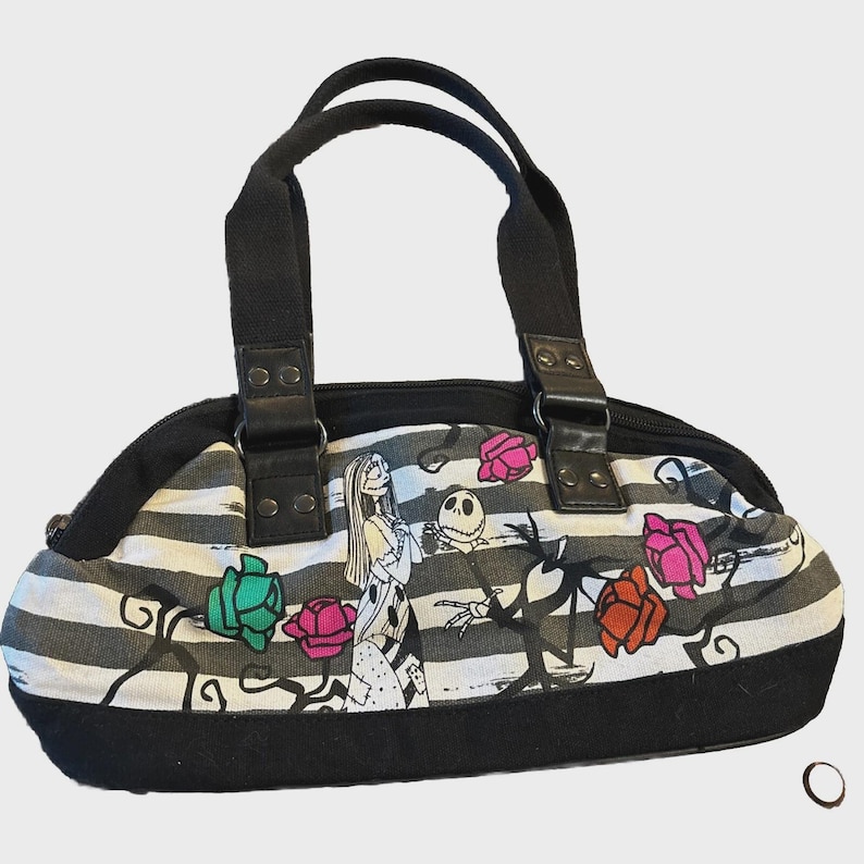 Puede incluir: Bolso de mano a rayas blancas y negras con personajes de The Nightmare Before Christmas. Tiene asas y ribetes negros, con cierre de cremallera. El dise&ntilde;o incluye a Sally, Jack Skellington y rosas de colores.