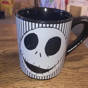 May include: Black and white ceramic mug featuring a smiling skull face design. The mug has a black interior and handle, with vertical black and white stripes on the exterior. A fun and unique coffee mug.