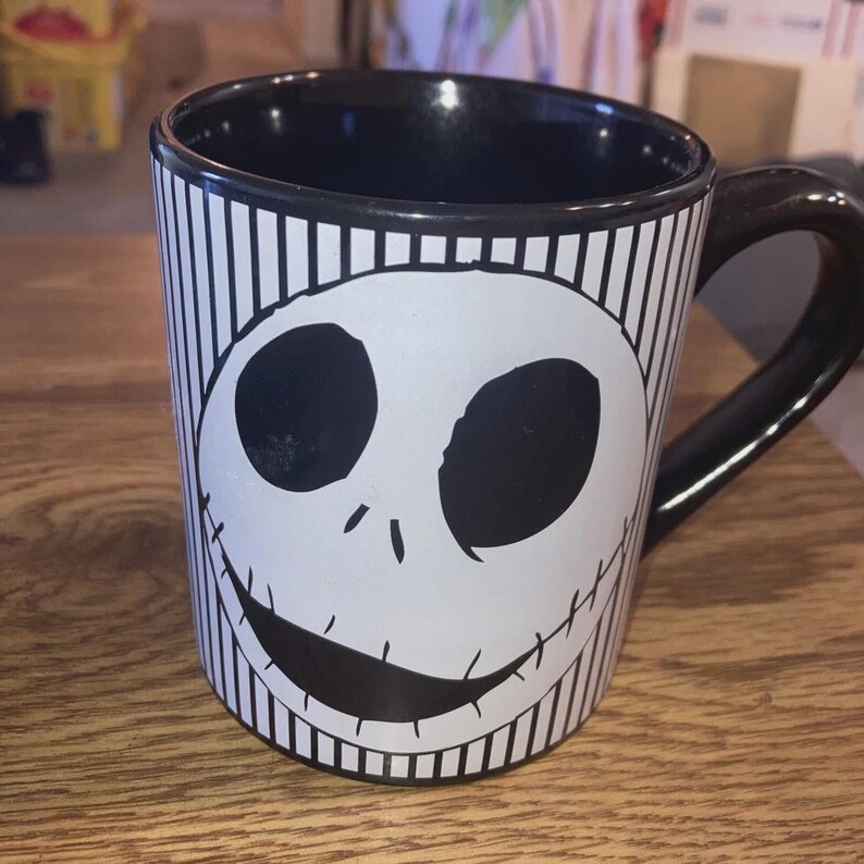 May include: Black and white ceramic mug featuring a smiling skull face design. The mug has a black interior and handle, with vertical black and white stripes on the exterior. A fun and unique coffee mug.