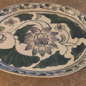 May include: Oval ceramic serving platter with a floral design. The off-white platter features a central lotus flower and leaves in blue and green. The rim is decorated with a blue floral border.