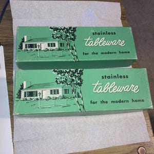 May include: Two vintage green boxes of stainless tableware. Each box features a white house illustration with a tree and the words "stainless tableware for the modern home" in white script.