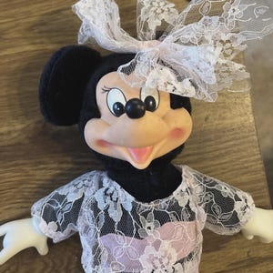 May include: A Minnie Mouse doll with a white lace bow, a black top, and a light purple dress. The doll has a pink face and a white lace top with short sleeves. The doll's arms are white.
