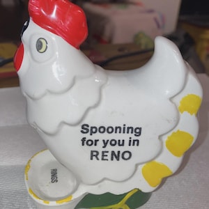 May include: A white ceramic hen-shaped spoon rest with a red comb and wattle, yellow accents, and a green base. The text "Spooning for you in RENO" is printed on the side. The spoon rest has a small circular area for rings.