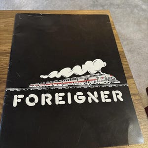 May include: Black album cover featuring the band name "FOREIGNER" in white block letters. Above the name is a stylized train with red and white accents, and a cloud of white smoke. The album cover is on a wooden surface.