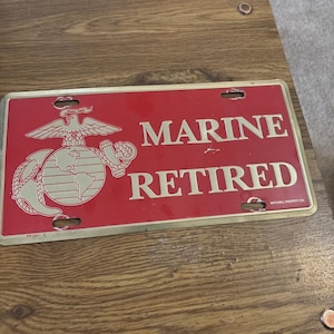 May include: A red metal license plate with the words "MARINE RETIRED" in gold lettering. The plate features the emblem of the United States Marine Corps. The plate has four mounting holes.