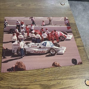 May include: A photograph of a white and red Indy race car with the number 2 and True Value branding on the side. The car is parked on a track with a pit crew in white and red uniforms standing nearby. A blue and white umbrella is attached to the car.