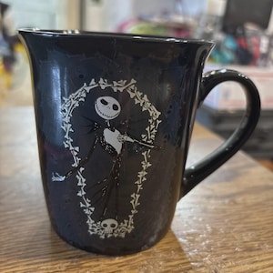 May include: Black ceramic mug featuring Jack Skellington from The Nightmare Before Christmas. The white graphic shows Jack inside a coffin-shaped frame with bones and a skull at the bottom. The mug has a curved handle.