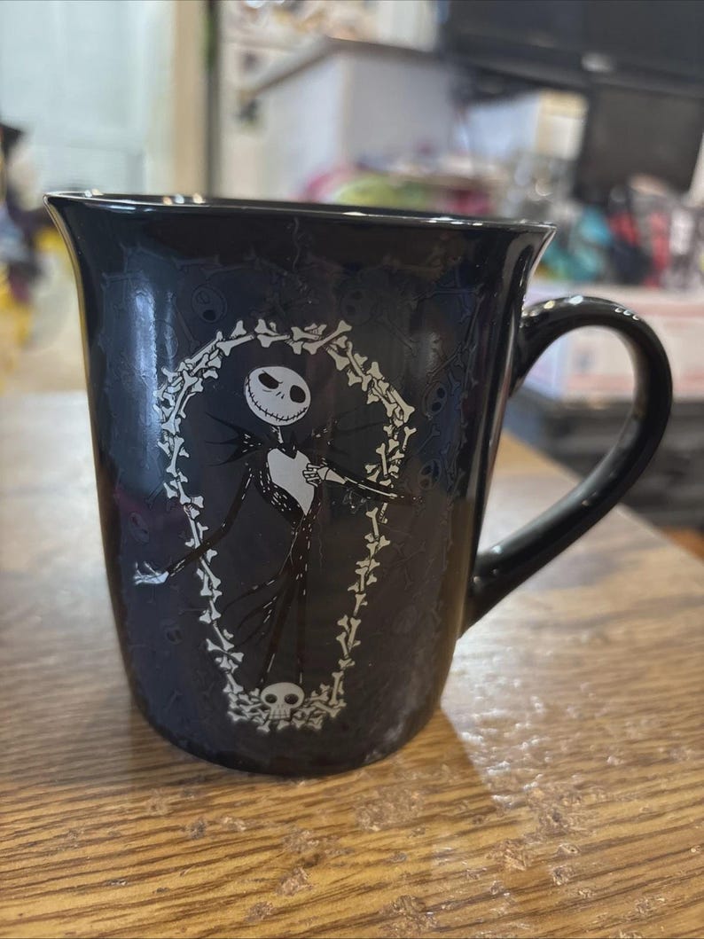 May include: Black ceramic mug featuring Jack Skellington from The Nightmare Before Christmas. The white graphic shows Jack inside a coffin-shaped frame with bones and a skull at the bottom. The mug has a curved handle.