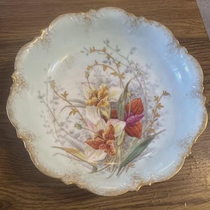 May include: A decorative, scalloped-edge bowl with a light blue interior and gold trim. The bowl features a floral design with orange, white, and yellow flowers, along with green leaves and delicate vines. The bowl is likely made of porcelain.