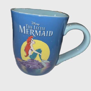 May include: A blue ceramic mug featuring the Disney movie "The Little Mermaid." The mug has a light blue handle and interior. The front of the mug displays Ariel, the mermaid, with a yellow moon and the movie title.