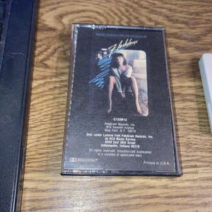 May include: A vintage cassette tape for the movie soundtrack "Flashdance." The cover features a woman in a seated pose with the title in a stylized font. The cassette case is clear with printed text and logos.
