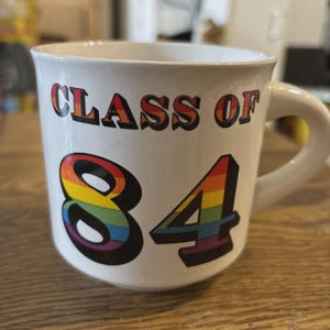 May include: White ceramic mug with the text "CLASS OF '84" in a rainbow gradient. The numbers "84" are also in a rainbow gradient. The mug has a curved handle and is sitting on a wooden surface.