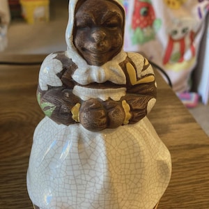 Puede incluir: Figura de cerámica vintage de una persona con rostro marrón y gorro blanco. La figura viste un vestido blanco con efecto craquelado. La figura tiene brazos marrones y una pajarita blanca. La base es marrón y amarilla.