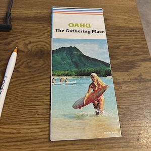 May include: Vintage travel brochure for Oahu, Hawaii, featuring a woman with a surfboard in the ocean. The brochure has the text "Oahu The Gathering Place" and a scenic image of a mountain and beach.