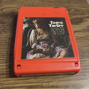 May include: A vintage red 8-track tape featuring Tanya Tucker's album, "Would You Lay With Me (In A Field of Stone)." The album art shows a woman in a dress. The tape is on a wooden surface.