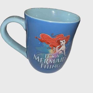 May include: Blue ceramic mug featuring Ariel from The Little Mermaid with red hair and a mermaid tail. The mug has a light blue handle and interior, with the text "It's a Mermaid Thing".