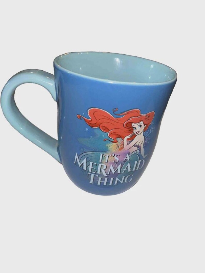 May include: Blue ceramic mug featuring Ariel from The Little Mermaid with red hair and a mermaid tail. The mug has a light blue handle and interior, with the text "It's a Mermaid Thing".