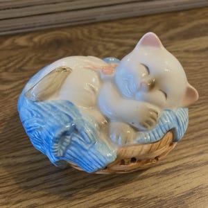 Puede incluir: Figura de cerámica de un gatito blanco durmiendo, acurrucado sobre una bola de lana azul. El gatito tiene orejas rosas y descansa en una cesta tejida. La figura está pintada con detalles brillantes.