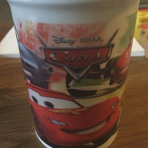 May include: White ceramic mug featuring characters from the Disney Pixar Cars film. The mug has a wraparound design with Lightning McQueen and other race cars. The Disney Pixar Cars logo is also visible.