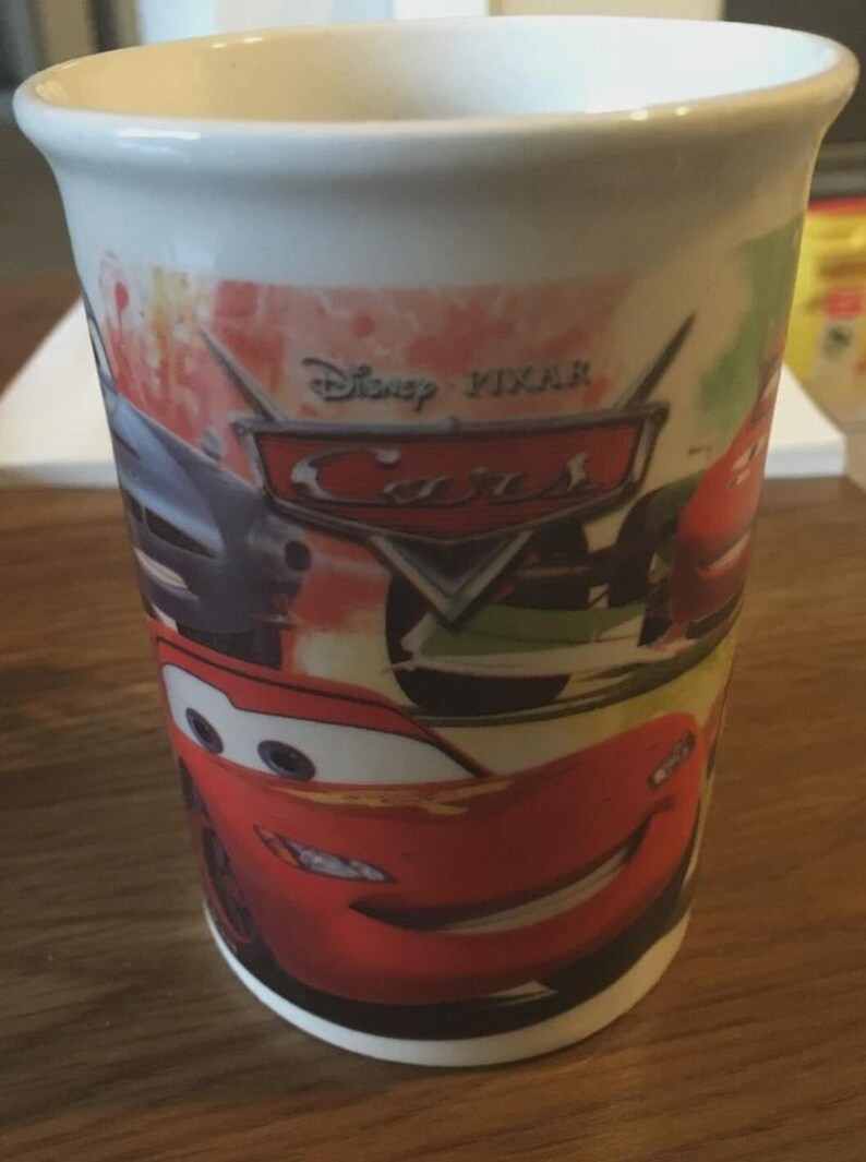 May include: White ceramic mug featuring characters from the Disney Pixar Cars film. The mug has a wraparound design with Lightning McQueen and other race cars. The Disney Pixar Cars logo is also visible.