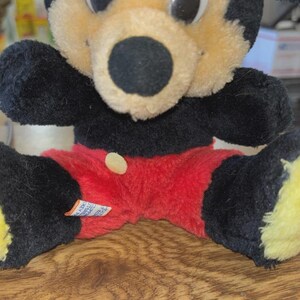 May include: A vintage Mickey Mouse plush toy. The plush features a black body, ears, and arms, with a tan face and red shorts. The toy has large white eyes and a black nose. A small tag is visible on the shorts.