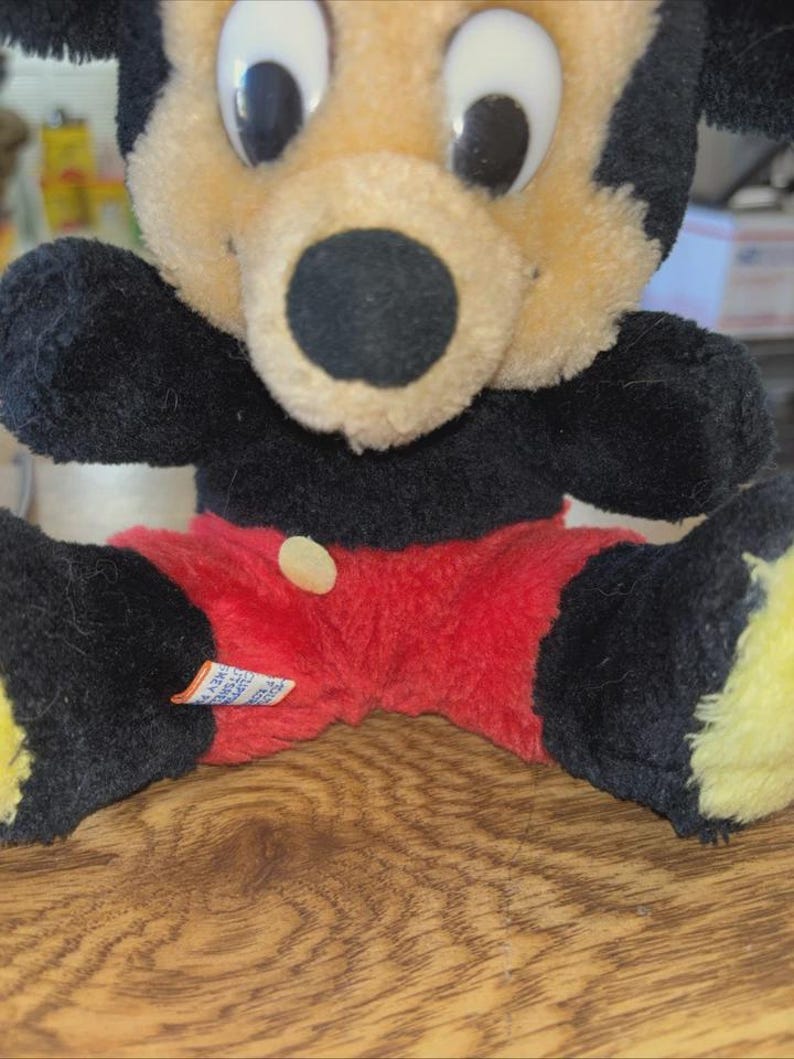 May include: A vintage Mickey Mouse plush toy. The plush features a black body, ears, and arms, with a tan face and red shorts. The toy has large white eyes and a black nose. A small tag is visible on the shorts.