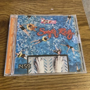 May include: A CD case featuring the band Sugar Ray's album cover. The cover art depicts people swimming in a pool with the band's name in red script. The time 14:59 is displayed on a digital clock.