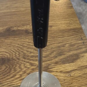 May include: A kitchen utensil comprising a black cylindrical handle connected to a slender metal rod and a flat circular metal base. The handle features the text "PAT. PENDING" embossed on its side, with further small text on the metal base.