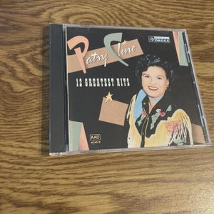 May include: A Patsy Cline "12 Greatest Hits" CD album in a clear plastic case. The album cover features a portrait of Patsy Cline with the album title in a stylized font. The background is black with a star and a Decca Records logo.