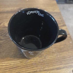 May include: Black ceramic mug with the text "The Pumpkin King" in white script. The mug has a rounded shape with a handle and is set against a wooden surface.
