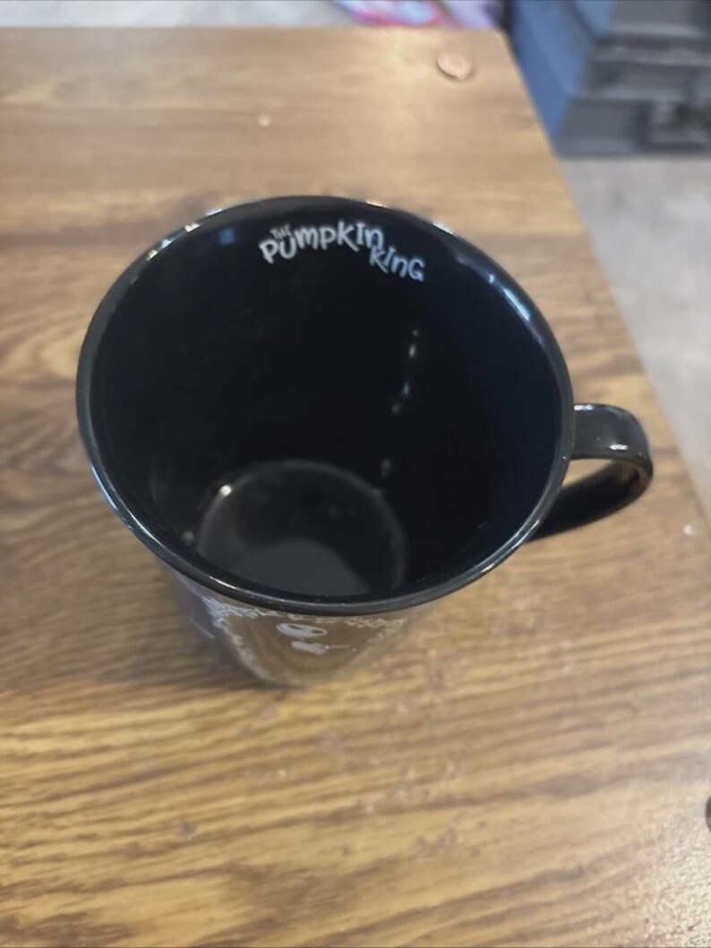May include: Black ceramic mug with the text "The Pumpkin King" in white script. The mug has a rounded shape with a handle and is set against a wooden surface.