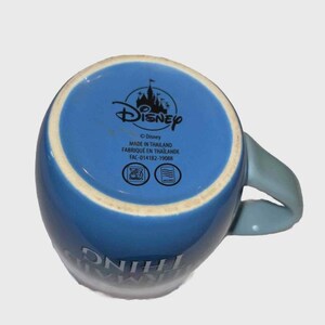 May include: Blue ceramic Disney mug with a light blue handle. The bottom of the mug features the Disney logo and text that reads "MADE IN THAILAND". The mug has the words "SOMETHING" and "MAGIC" printed on the side.