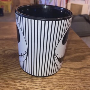 May include: Black and white striped ceramic mug featuring a design of Jack Skellington's face from The Nightmare Before Christmas. The mug has a black interior and a cylindrical shape, perfect for hot beverages.