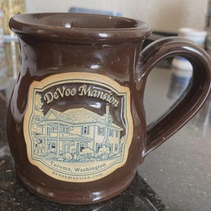 May include: A white ceramic mug with a black handle and the text "But First, Coffee" in a black, handwritten-style font. The mug is on a white surface.