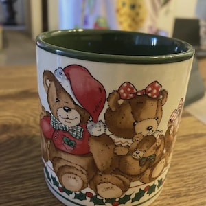Op de afbeelding: Een keramische mok met een witte buitenkant en een donkergroene binnenrand. De mok is voorzien van een kleurrijke illustratie van drie bruine teddyberen, waarvan er één een rode kerstmuts en een groen geruite vlinderdas draagt.