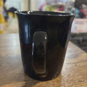 May include: A glossy black coffee mug with a curved handle. The mug has a slightly flared rim and sits on a wooden surface. The mug is empty and ready to be filled with a hot beverage.