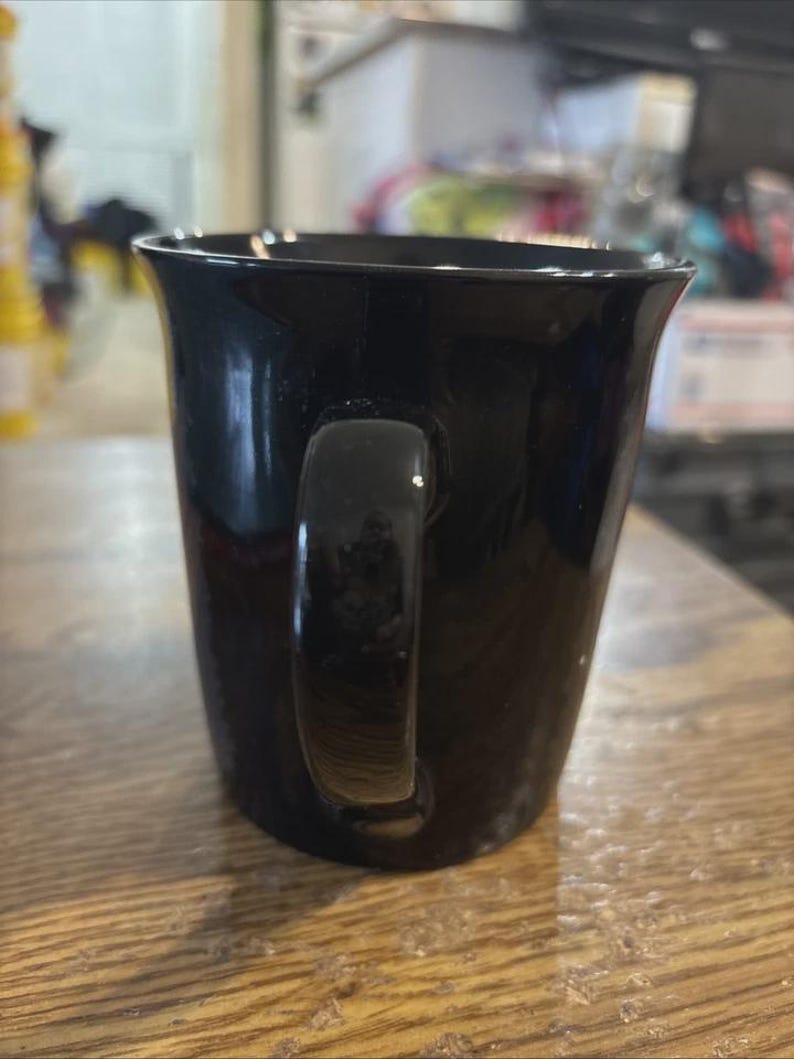 May include: A glossy black coffee mug with a curved handle. The mug has a slightly flared rim and sits on a wooden surface. The mug is empty and ready to be filled with a hot beverage.