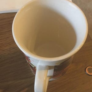 May include: White ceramic coffee mug with a curved handle. The interior is a light beige colour. The exterior features a design with red, black, and brown accents. The mug is on a wooden surface.