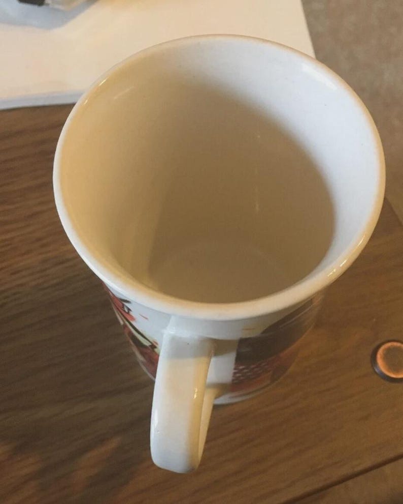 May include: White ceramic coffee mug with a curved handle. The interior is a light beige colour. The exterior features a design with red, black, and brown accents. The mug is on a wooden surface.