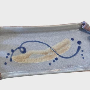 May include: Rectangular ceramic serving tray with a light blue-gray base. It features a tan and blue abstract design and brown, looped handles. The tray is suitable for serving appetizers or displaying decorative items.