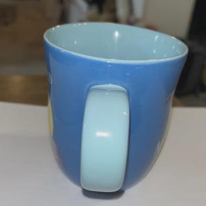May include: A blue ceramic mug with a light blue interior and handle. The mug has a curved shape and a glossy finish. The handle is a light blue color. The mug appears to have some text and designs on the exterior.
