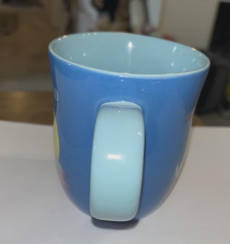 May include: A blue ceramic mug with a light blue interior and handle. The mug has a curved shape and a glossy finish. The handle is a light blue color. The mug appears to have some text and designs on the exterior.