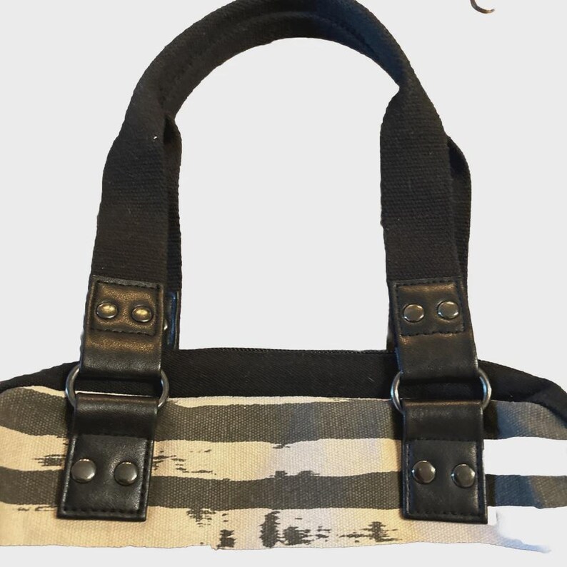 Puede incluir: Un bolso tote de lona con rayas horizontales negras y grises. El bolso tiene asas negras y detalles de cuero negro. Las asas est&aacute;n unidas con anillos y remaches de metal plateado.