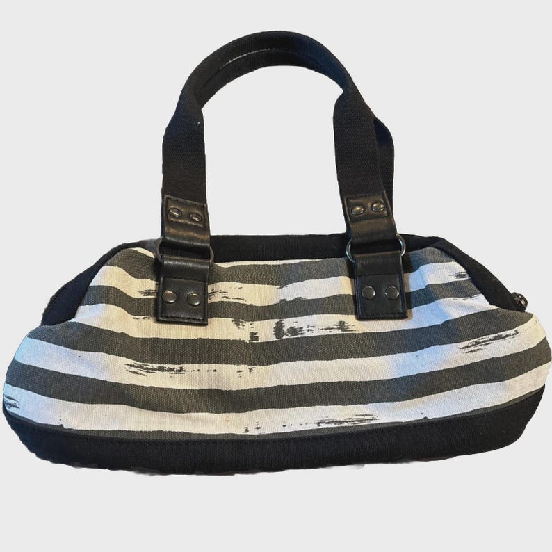 Puede incluir: Bolso de mano a rayas blancas y negras con asas y ribetes negros. El bolso tiene forma rectangular con extremos redondeados y est&aacute; hecho de un material similar a la lona. Las asas est&aacute;n sujetas con correas de cuero negro.