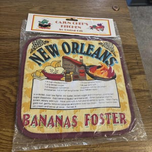 May include: A square fabric pot holder in a clear plastic package with a header card labelled Cajun Chef's Kitchen. The pot holder features a yellow background with banana illustrations, the text New Orleans Bananas Foster, and a recipe.